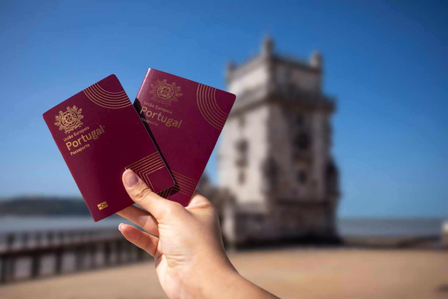 Two Portuguese passports held in front of the iconic Belém Tower (Torre de Belém) in Lisbon, Portugal.